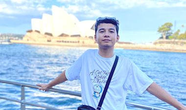A young man in a white T-shirt stands by a waterfront railing, with the Sydney Opera House in the background under a blue sky.