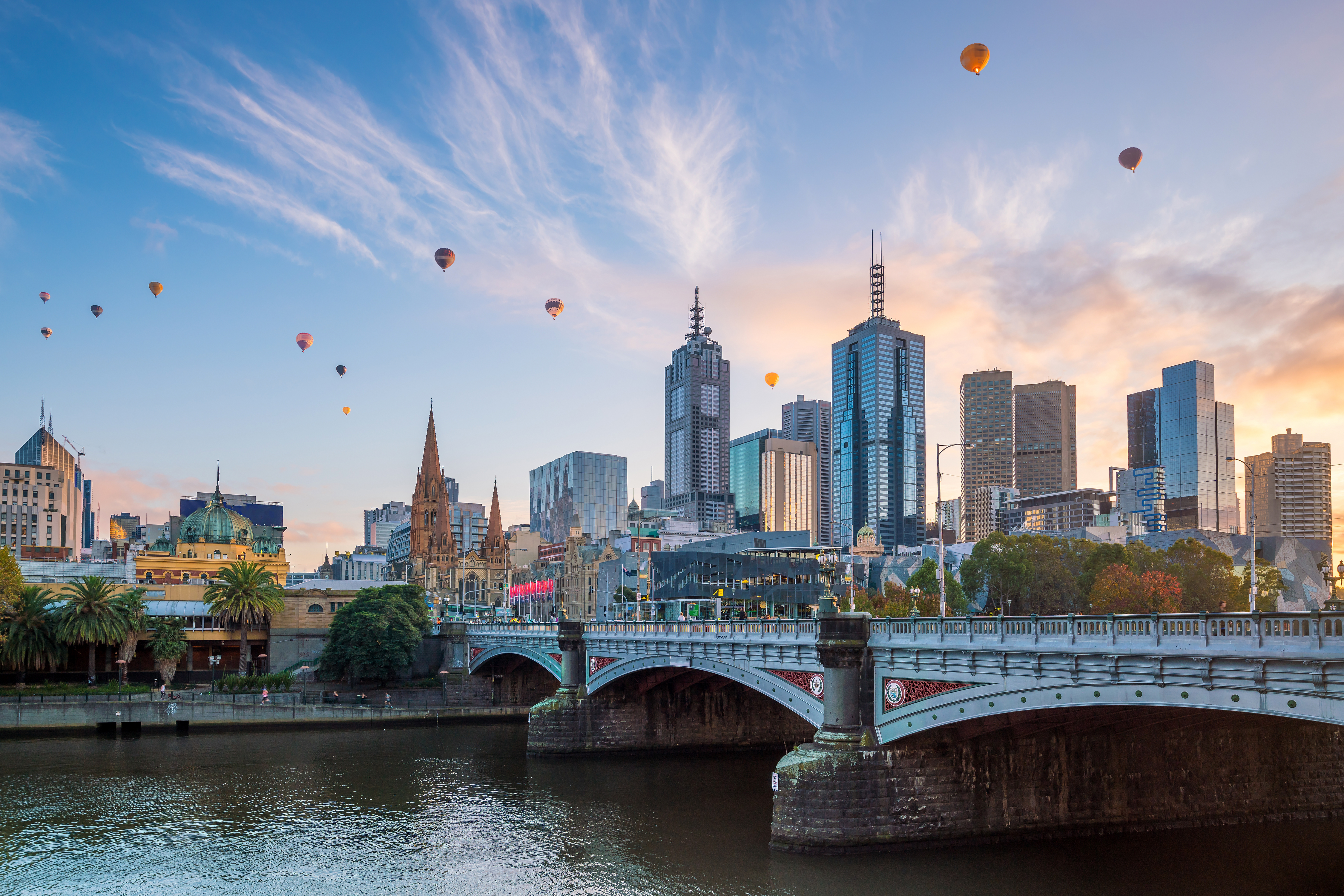Hot air balloons float over Melbourne's skyline at sunset, with the Yarra River and Princes Bridge in the foreground