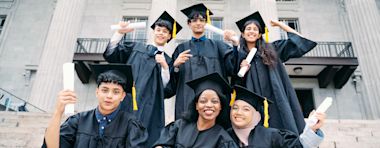 Six graduates in caps and gowns stand on steps, smiling and holding diplomas, with a building featuring large columns in the background.