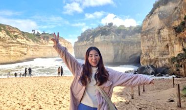A young woman smiles and spreads her arms wide on a sandy beach with rocky cliffs and blue sky in the background. Several people are seen walking near the shoreline and ocean waves.