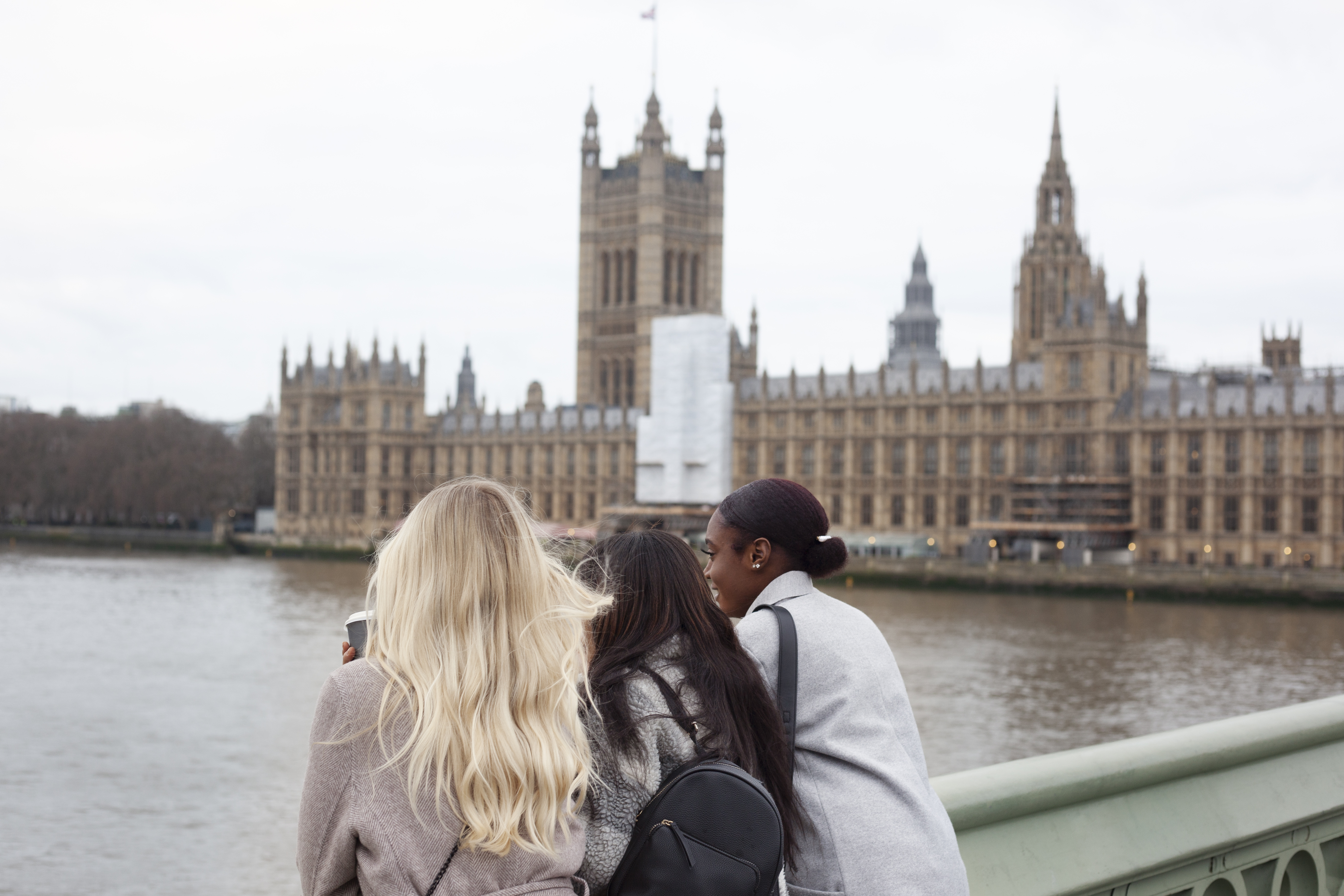 Three women stand on a bridge looking across the River Thames at the Houses of Parliament in London, with the iconic clock tower (Big Ben) in the background on a cloudy day.