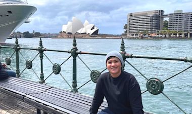 A person wearing a gray beanie and dark sweater sits on a bench by the waterfront, smiling, with the Sydney Opera House and modern buildings visible across the water under a cloudy sky.