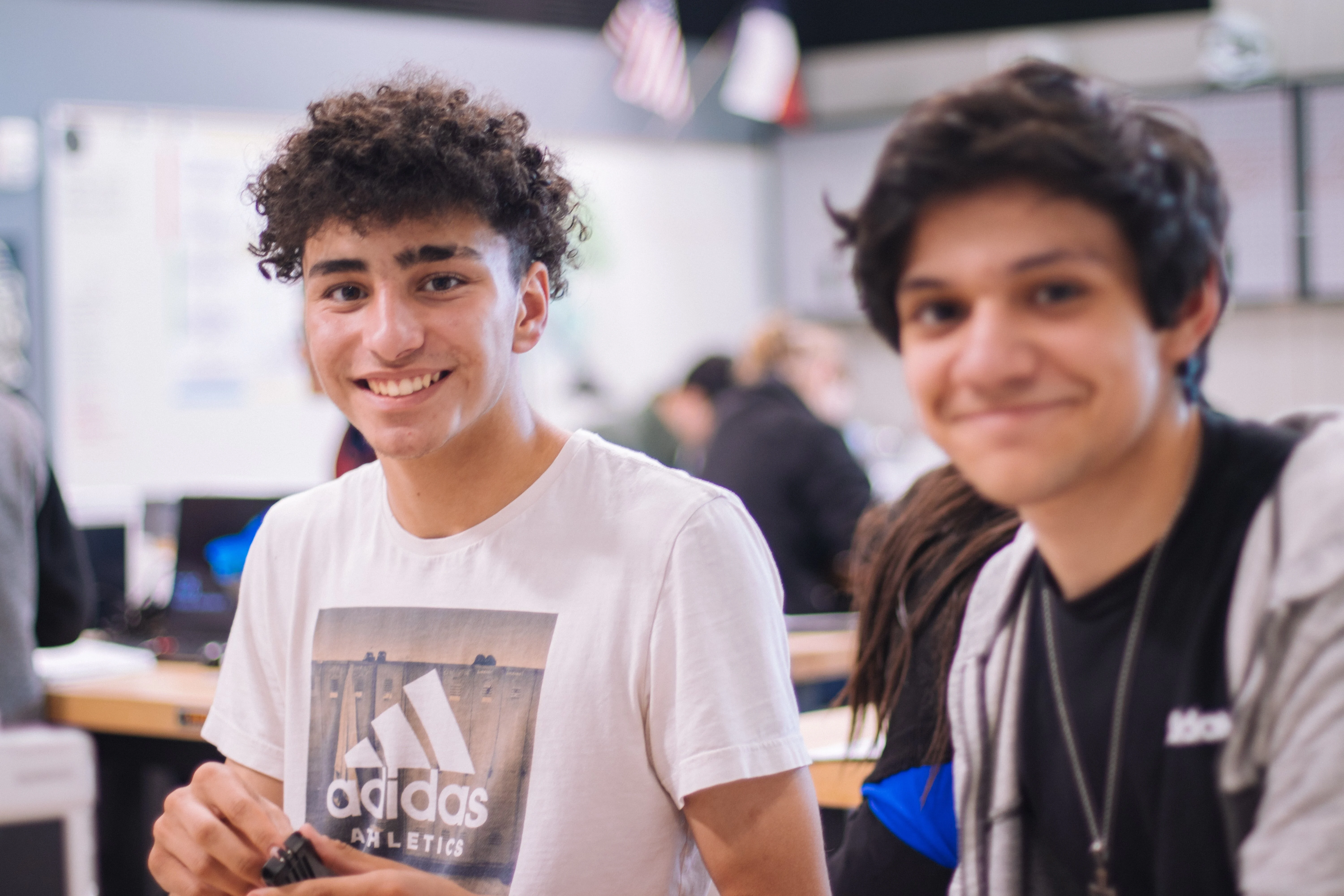 Two smiling students in a classroom, one wearing a white Adidas t-shirt and the other in dark casual wear, with computers visible behind them.