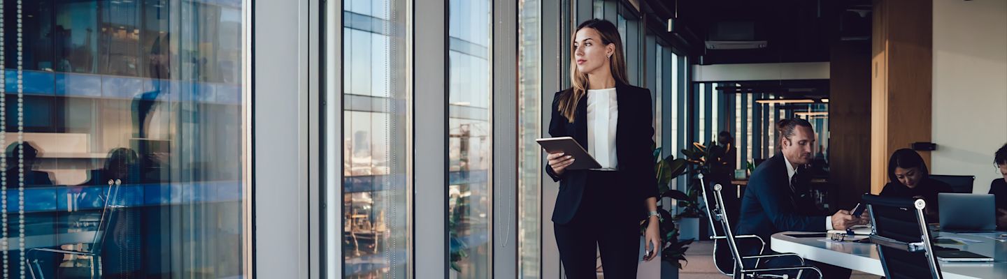 Professional in black suit holding tablet stands by office windows while colleagues work at desks in modern workspace with city views