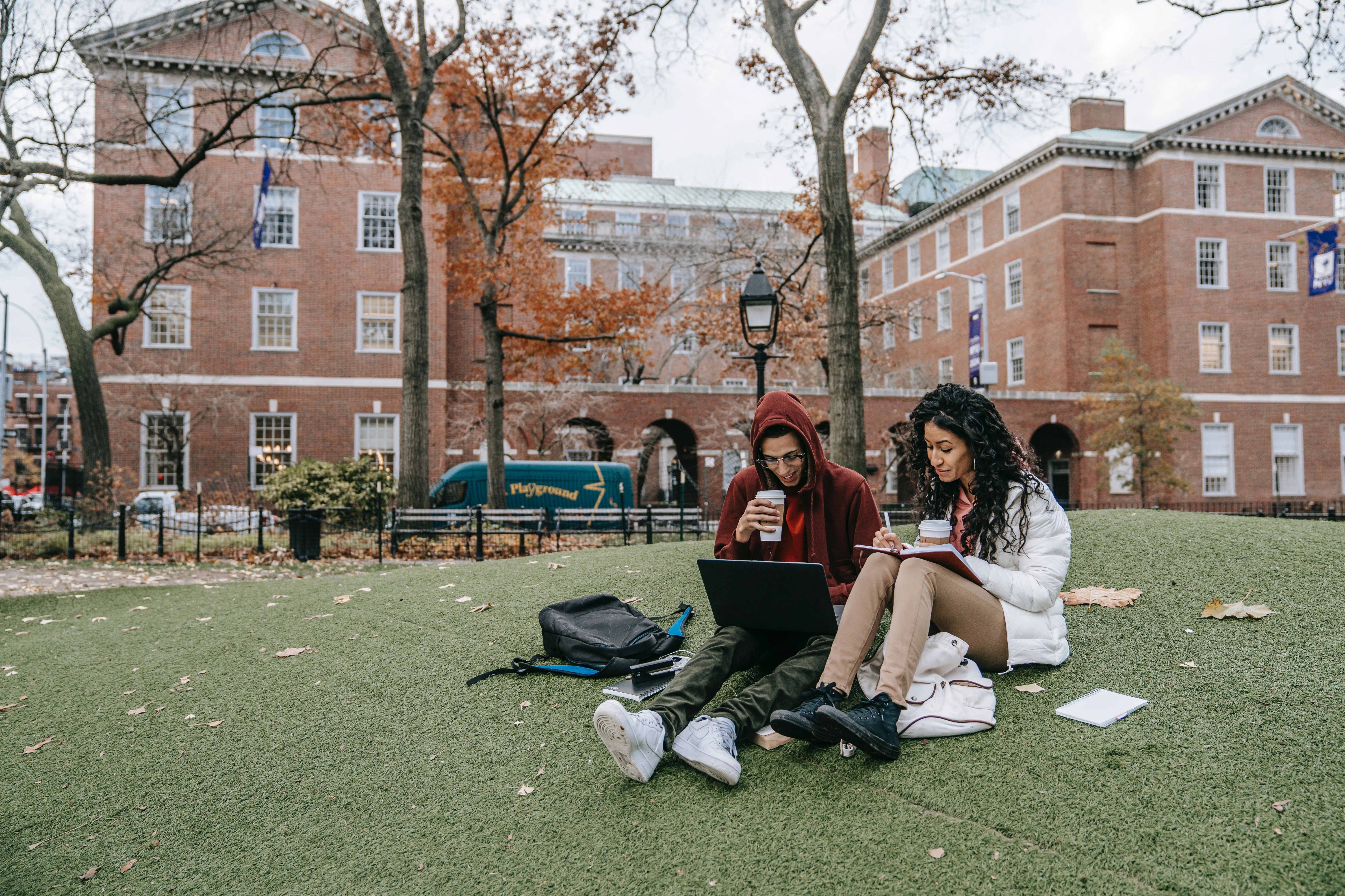 Students studying on a lawn in front of a historic brick university building during autumn, with fallen leaves scattered around