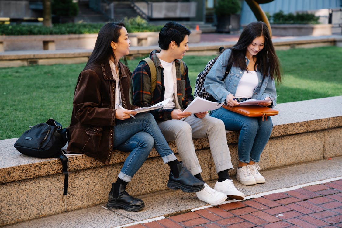 There are three students sitting on a ledge. They are smiling while looking at their notes. 