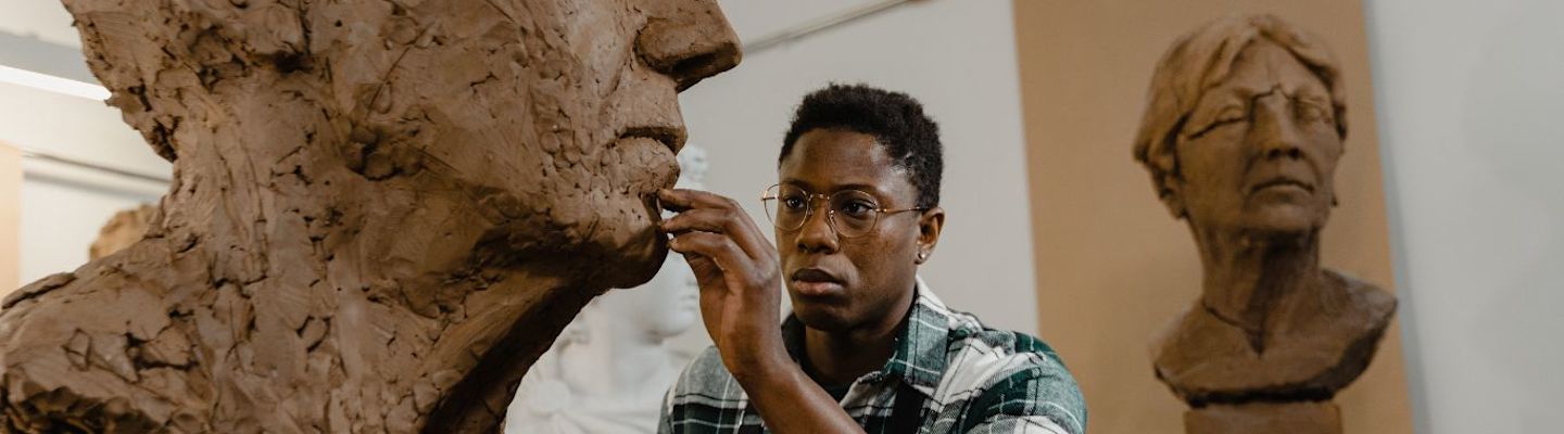 A person wearing glasses and a plaid shirt sculpts a large, detailed clay bust in an art studio. Another clay bust of a different person is visible in the background.