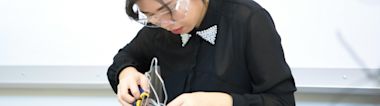 A person wearing safety goggles and a black shirt with a beaded collar is focused on working with electronic components and tools at a desk.