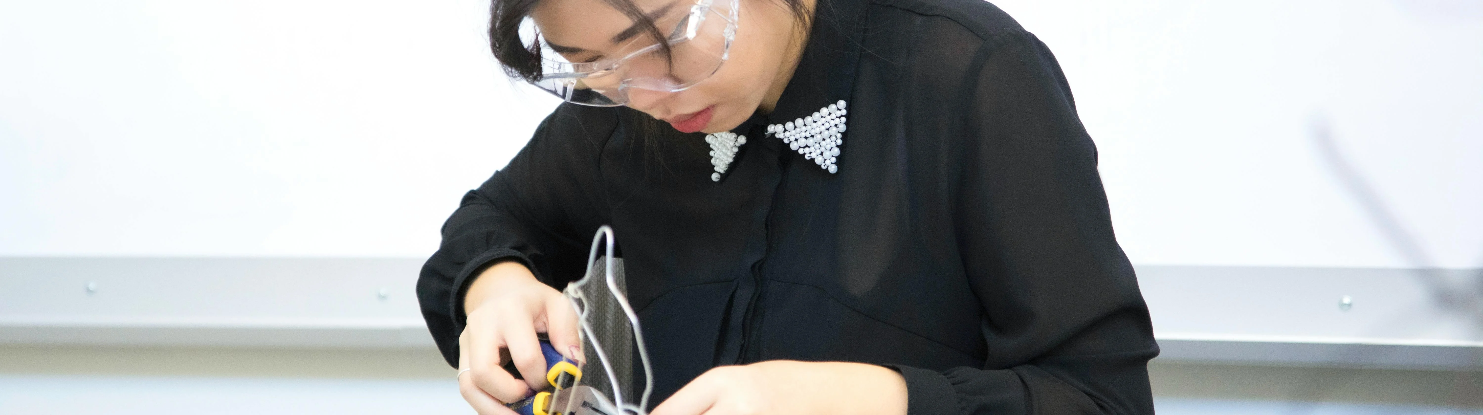 A person wearing safety goggles and a black shirt with a beaded collar is focused on working with electronic components and tools at a desk.