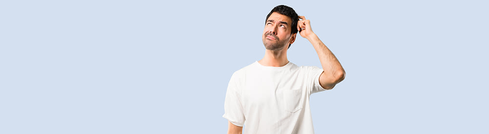Young man with white shirt having doubts and with confuse face expression while scratching head on isolated blue background