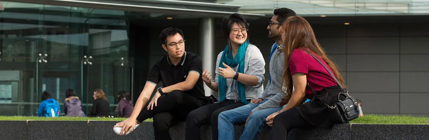 Four young adults sit on a low wall outside a modern building, talking and smiling together. Three people face one person who is gesturing while speaking. All are casually dressed.