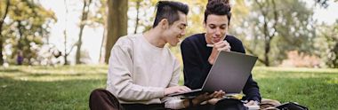 Two young adults sit on grass in a park, smiling and working together on a laptop. One holds a pen while the other types, with notebooks and trees visible in the background.