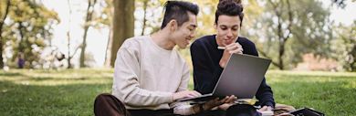 Two young adults sit on grass in a park, smiling and working together on a laptop. One holds a pen while the other types, with notebooks and trees visible in the background.
