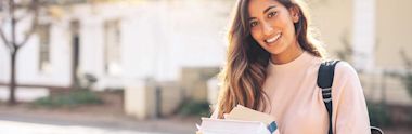 A young woman with long hair, wearing a light-colored sweater and backpack, smiles while holding several books outdoors on a sunny day.