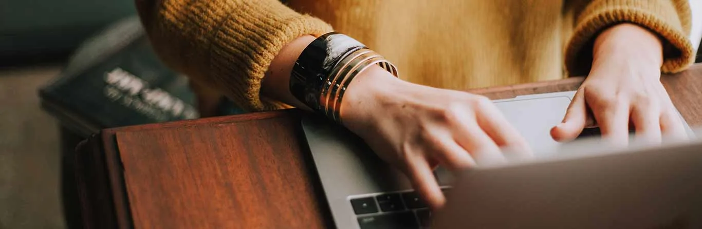 A person wearing a yellow sweater and bracelets types on a laptop at a wooden desk. A book is partially visible in the background.