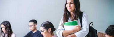 A young woman holding books and wearing a backpack stands and smiles in a classroom, while other students sit at desks, focused on their work.