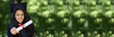 Graduate in cap and gown holds up diploma while smiling outdoors against blurred green foliage background