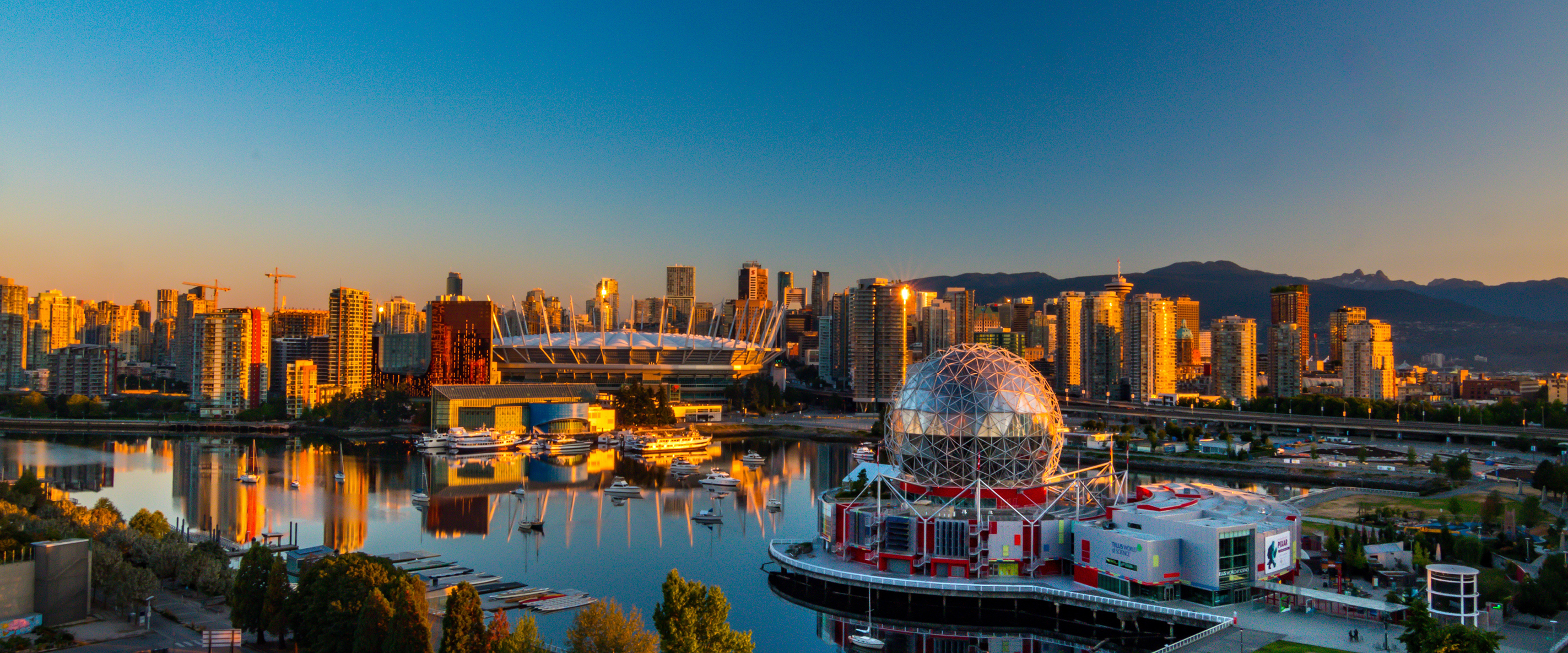 Panoramic view of Vancouver skyline at sunset, featuring high-rise buildings, a large stadium, and the geodesic dome of Science World reflected in calm water with mountains in the background.
