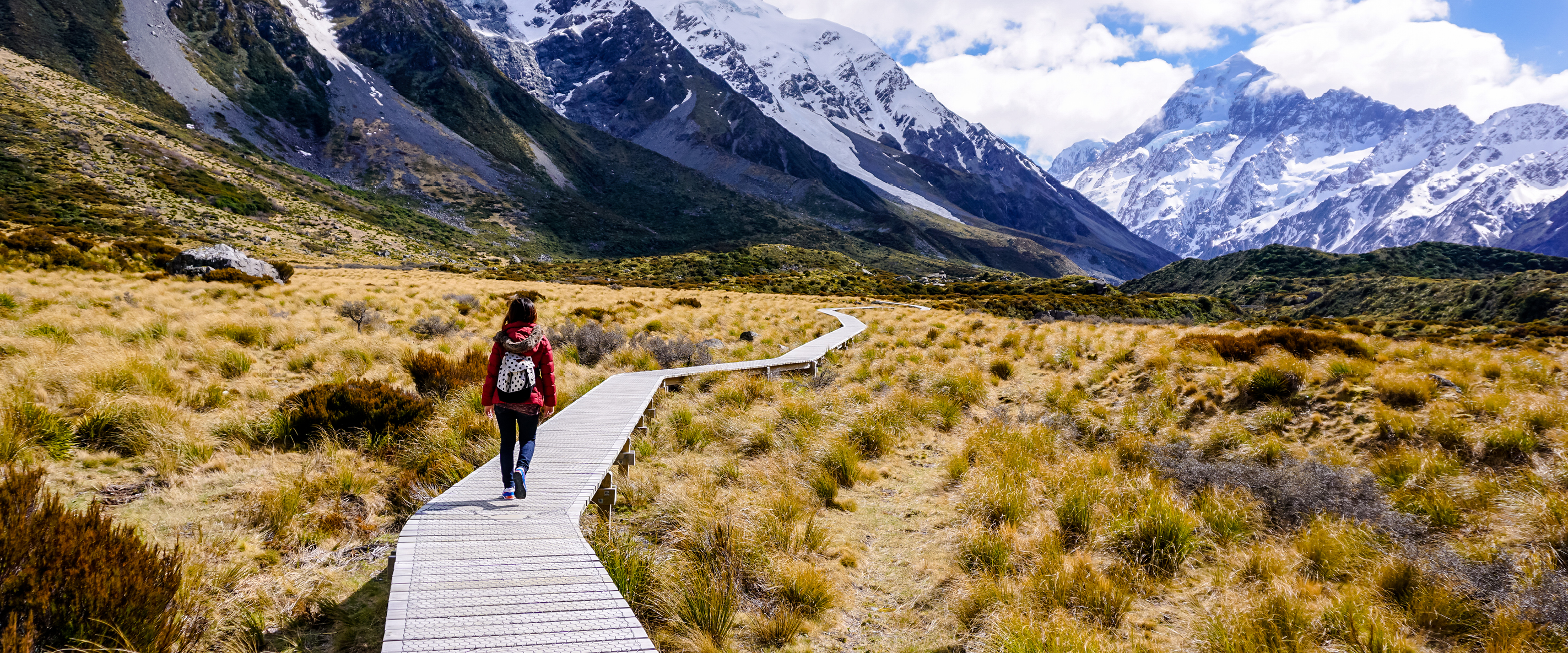 A person with a backpack walks on a wooden path through grassy fields toward snow-capped mountains under a partly cloudy sky.