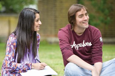 Two people sitting on grass outside, smiling and talking. One holds a notebook and pen, while the other wears a maroon sweatshirt. A blurred brick wall and greenery are in the background.