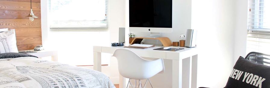 A modern bedroom with a workspace featuring a white desk and an Apple computer. A comfortable chair is placed in front, and theres a bed with a city-themed duvet. A New York pillow is on a nearby chair. Natural light filters through the window blinds.