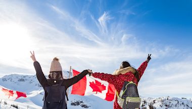 Two people in winter clothing stand with raised arms, holding Canadian flags. They face a snowy mountain landscape under a bright blue sky.