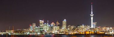 A nighttime view of Auckland’s city skyline, featuring illuminated high-rise buildings and the iconic Sky Tower, with reflections on the harbor water in the foreground.
