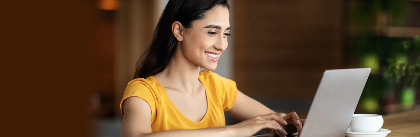 A young woman wearing a yellow shirt is smiling while using a laptop at a table. A white cup and saucer are on the table, and the background is softly blurred.