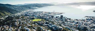 Aerial view of Wellington, New Zealand, showing city buildings, a port, and surrounding hills next to a large body of water under a clear sky.
