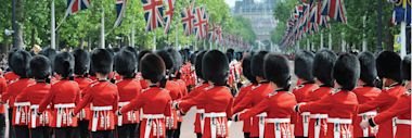 Soldiers in red uniforms and tall black hats march in formation on a tree-lined street decorated with British flags, likely during a ceremonial parade in the UK.