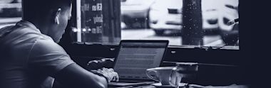 Person working on laptop at cafe window seat with coffee cup, captured in black and white monochrome