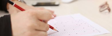 A person holds a red pen and fills in bubbles on a standardized test answer sheet on a desk, with a smartphone and eyeglasses in the background.