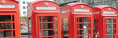 Four classic red British telephone booths stand in a row on a city street, with buildings and another phone booth visible in the background.