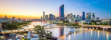 Brisbane city skyline at sunset with modern high-rise buildings, bridges over the Brisbane River, and reflections on the water, under a clear sky.