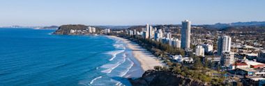 Aerial view of a coastal city with high-rise buildings along a sandy beach, blue ocean waves, and tree-lined streets; green hills and more coastline visible in the background.