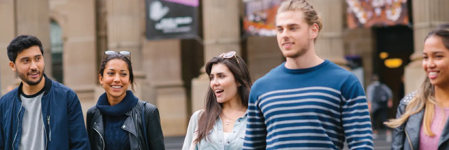 A group of five young adults walk together outdoors in an urban setting, smiling and talking, with buildings and banners visible in the background.