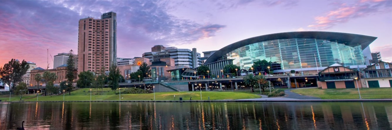 A cityscape at sunset with modern and historic buildings, a large glass-walled convention center, green lawns, trees, and a calm river reflecting the skyline.