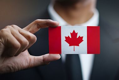 A person wearing a suit holds a small Canadian flag card between their fingers. The focus is on the card, which features a red maple leaf in the center with red bars on each side.