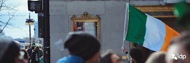 A crowd gathers near a building on Jackson Blvd; someone waves an Irish flag. A gold-framed mirror hangs on the wall, and the IDP logo appears in the corner.