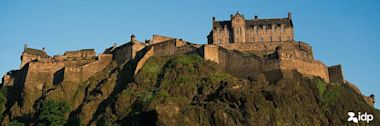 Edinburgh Castle perched atop a rocky cliff against blue sky, with stone fortifications and buildings crowning the hilltop