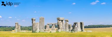 Stonehenge on a grassy plain under a blue sky with scattered clouds. The ancient stone structure stands prominently, with fields and trees in the background. The IDP logo appears in the top left corner.
