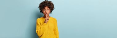 Person in bright yellow sweater making shushing gesture with finger to lips against light blue background, with curly natural hair