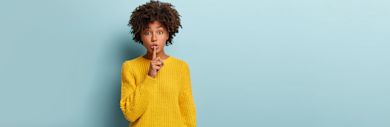 Person in bright yellow sweater making shushing gesture with finger to lips against light blue background, with curly natural hair
