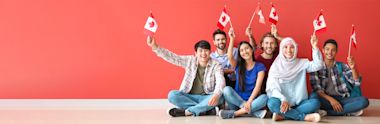 Diverse group of young people sitting on floor against coral wall, waving Canadian flags with enthusiasm and bright smiles