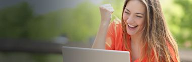 Excited person in coral-colored top celebrating while looking at laptop outdoors with blurred green background