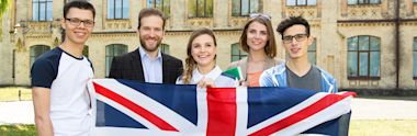 Group of students and instructor holding British flag outside historic university building with ornate brick architecture
