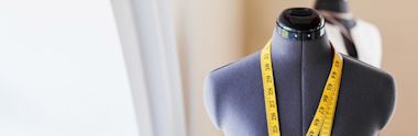 A dress form mannequin with a yellow measuring tape draped around its neck, set against a blurred background in a bright room.