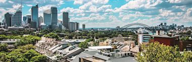 Sydney skyline view from residential area, showing Harbor Bridge, Opera House, and modern skyscrapers under blue sky with white clouds