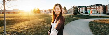 Person taking selfie on winding path through campus green at sunset, with residential buildings in background and warm golden light