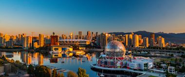 A panoramic view of Vancouver at sunset, featuring Science World’s geodesic dome in the foreground, tall city buildings, and reflections on the calm water under a clear blue sky.